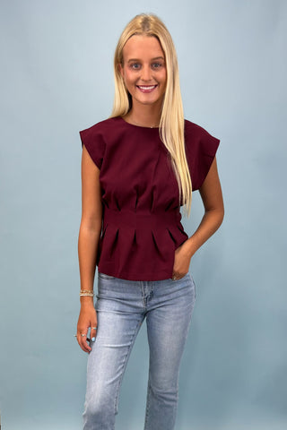 Woman wearing a burgundy peplum top and light blue jeans against a gray background