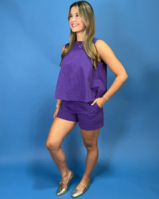 A woman modeling a matching purple sleeveless top and shorts set against a solid blue studio background.