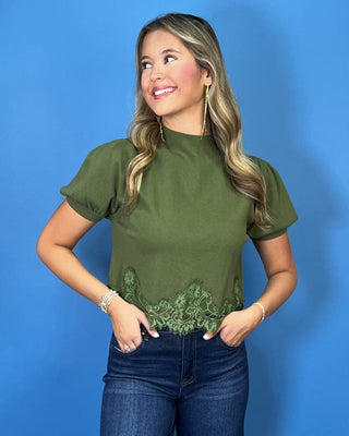 Olive green puff sleeve knit top with mock neckline and delicate lace hem detail, styled with dark wash jeans.