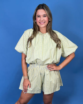 A woman modeling a light yellow and green polka dot romper with short puff sleeves and a matching waist tie, standing against a solid blue background.