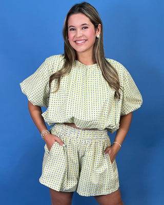 A woman modeling a light yellow two-piece set with a green and white polka dot pattern, featuring a short-sleeve top with puff sleeves and matching high-waisted shorts, set against a solid blue background.