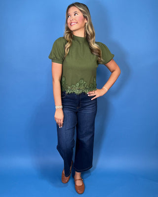 Olive green puff sleeve knit top with mock neckline and delicate lace hem detail, styled with dark wash jeans.
