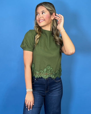 Olive green puff sleeve knit top with mock neckline and delicate lace hem detail, styled with dark wash jeans.