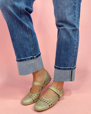 Gold flat shoes worn with rolled-up blue jeans on a pink background