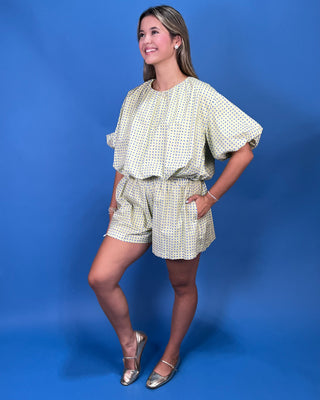 a light yellow and blue polka dot romper with short puff sleeves and a matching waist tie, standing against a solid blue background.