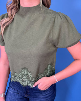 Olive green puff sleeve knit top with mock neckline and delicate lace hem detail, styled with dark wash jeans.