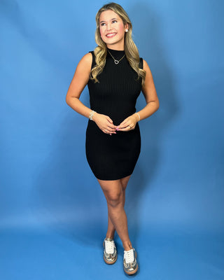 A young woman with blonde hair stands against a solid blue studio background, wearing a sleeveless black ribbed mock-neck mini dress