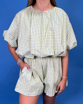 a light yellow and blue polka dot romper with short puff sleeves and a matching waist tie, standing against a solid blue background.
