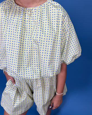 a light yellow and blue polka dot romper with short puff sleeves and a matching waist tie, standing against a solid blue background.