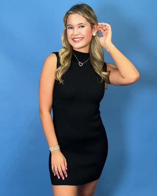 A woman with blonde hair wearing a sleeveless black ribbed mock neck mini dress, standing against a solid blue background.