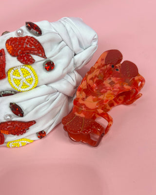 Hand holding a pair of white headband with red crawfish designs against a pink background