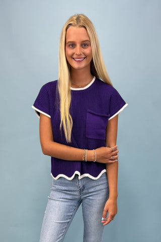 Woman wearing a purple top with white trim and blue jeans against a gray background
