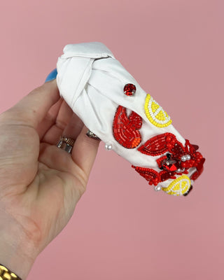 Hand holding a pair of white headband with red crawfish designs against a pink background