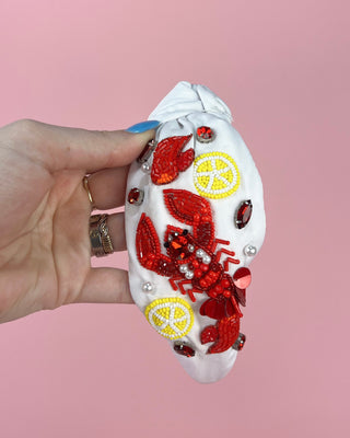 Hand holding a pair of white headband with red crawfish designs against a pink background