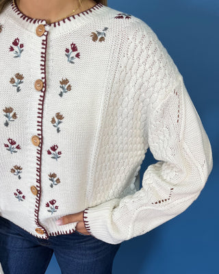 a cream cable-knit button-up sweater with embroidered floral details and brown buttons
