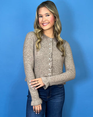 Taupe knit cardigan with delicate ruffle trim, pearl-style buttons, ruffled hem, and feminine flared cuffs.