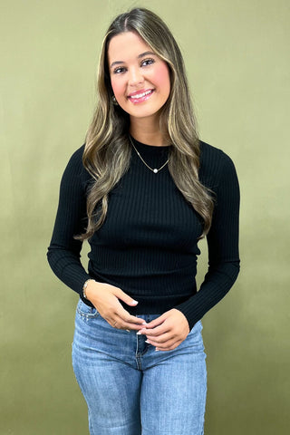 Woman wearing a black ribbed sweater and blue jeans against a green background