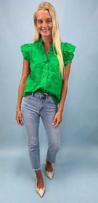 Woman wearing a green blouse with ruffled sleeves and blue jeans on a light gray background