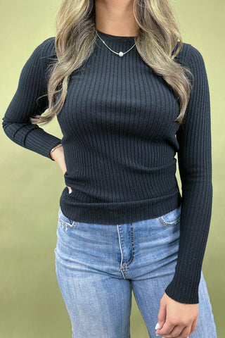 Person wearing a black ribbed sweater and blue jeans against a green background