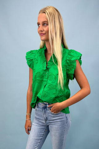 Woman wearing a green blouse with ruffled details and light blue jeans against a gray background