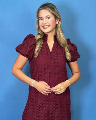 A smiling blonde woman wearing a short-sleeve burgundy eyelet dress with puff sleeves