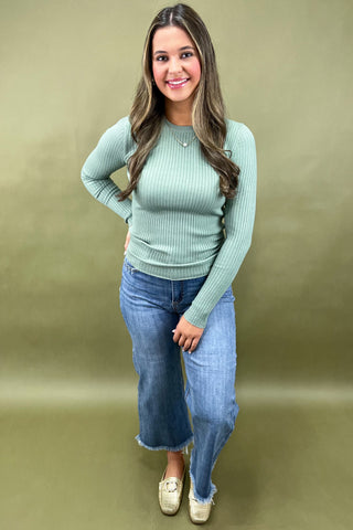 Woman wearing a green ribbed sweater and blue jeans against a beige background