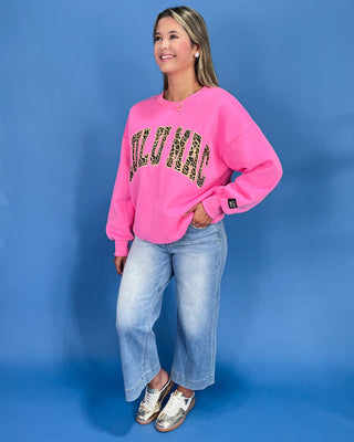 Woman wearing a pink sweatshirt with leopard print text and blue jeans against a blue background
