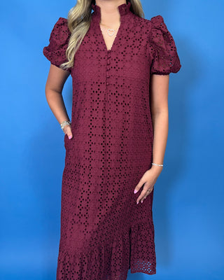 A woman wearing a burgundy eyelet lace midi dress with puff sleeves, a V-neckline, and side pockets, posed against a solid blue background.