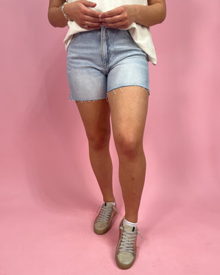 Person wearing light blue denim shorts and beige sneakers on a pink background