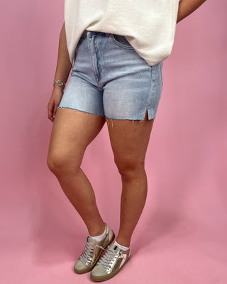 Person wearing light blue denim shorts and white sneakers on a pink background