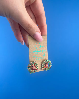 Shop the Cardinal In Wreath Earrings at All About You Boutique! Festive red cardinal studs with green Christmas wreaths and sparkling red berry accents.