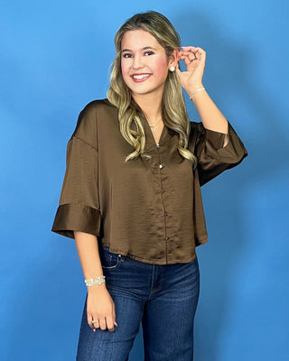 A woman modeling a brown satin-like blouse with wide sleeves and front buttons, paired with dark blue wide-leg jeans and white sneakers