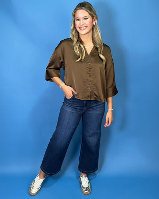 A woman modeling a brown satin button-up blouse with three-quarter sleeves, paired with dark blue wide-leg jeans and white sneakers