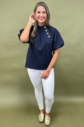 Woman wearing a navy blouse with puff sleeves and white pants against a green background