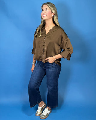 A woman modeling a brown satin-like button-up blouse with three-quarter sleeves and blue jeans
