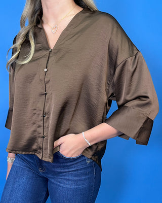 A woman modeling a brown satin button-up blouse with three-quarter sleeves, paired with dark blue wide-leg jeans and white sneakers