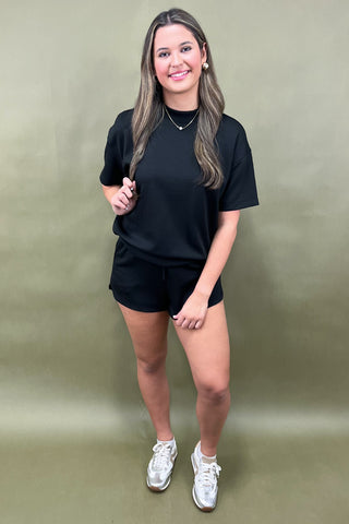 Woman wearing a black t-shirt and shorts with white sneakers against a green background