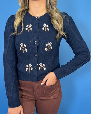 Navy cable knit cardigan with embroidered white daisies, button front, crew neckline, and long sleeves.