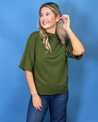 A woman with blonde hair wearing an olive green short-sleeve mock neck top and blue jeans