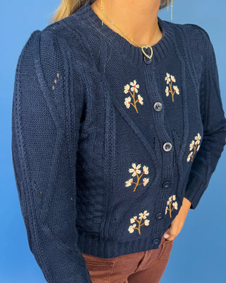 Navy cable knit cardigan with embroidered white daisies, button front, crew neckline, and long sleeves.