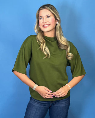 A woman with blonde hair wearing an olive green short-sleeve mock neck top and blue jeans