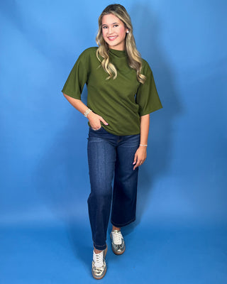 A woman with blonde hair wearing an olive green short-sleeve mock neck top and blue jeans