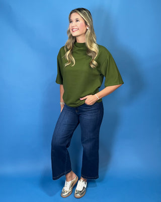 A woman modeling a short-sleeve olive green mock neck top with dark blue jeans and white sneakers