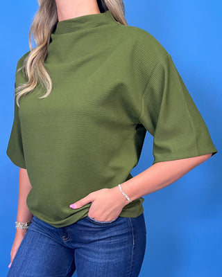 A woman wearing a textured olive green short-sleeve mock neck top and blue jeans