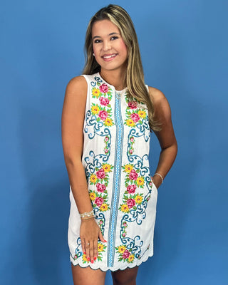 Woman wearing a white sleeveless dress with colorful floral embroidery against a blue background