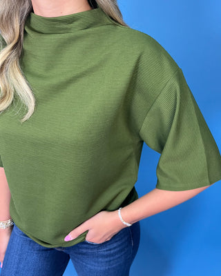 A woman wearing a ribbed olive green mock neck top with elbow-length sleeves, paired with blue jeans