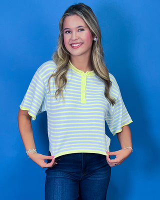 A young woman with blonde hair is wearing a short-sleeve striped henley shirt with neon yellow and light blue horizontal stripes, paired with dark blue jeans