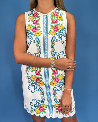 Woman wearing a white sleeveless dress with colorful floral embroidery against a blue background