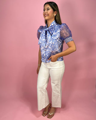 Woman wearing a blue floral blouse with a large bow and white pants on a pink background