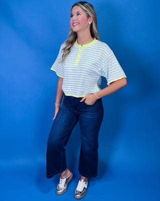 A woman modeling a short-sleeve striped henley top with neon yellow trim, paired with dark blue jeans and metallic sneakers